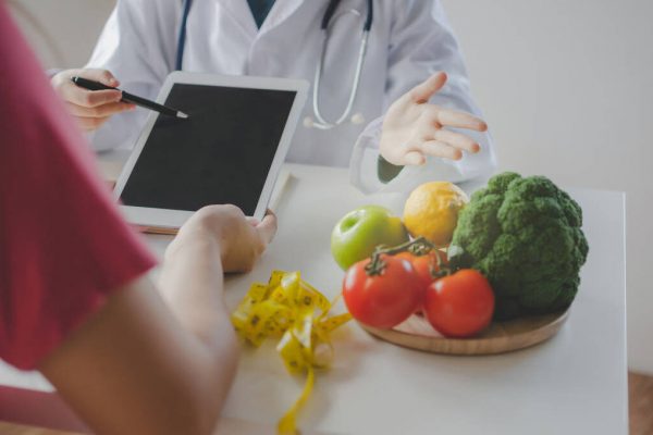 friendly nutritionist female doctor or medical using tablet and talking about diet plan with patient on desk at laboratory room at office hospital, food science, healthy food and dieting concept